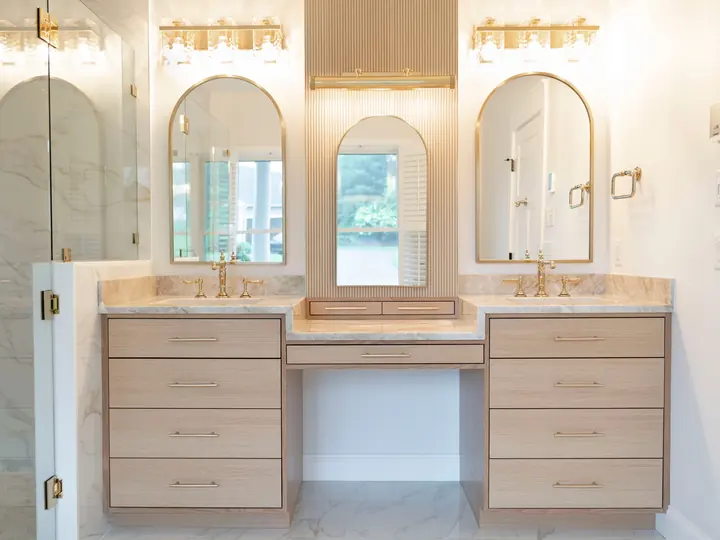 Bright bathroom with two vanities and a makeup counter in white stone, arched mirrors and brass lights
